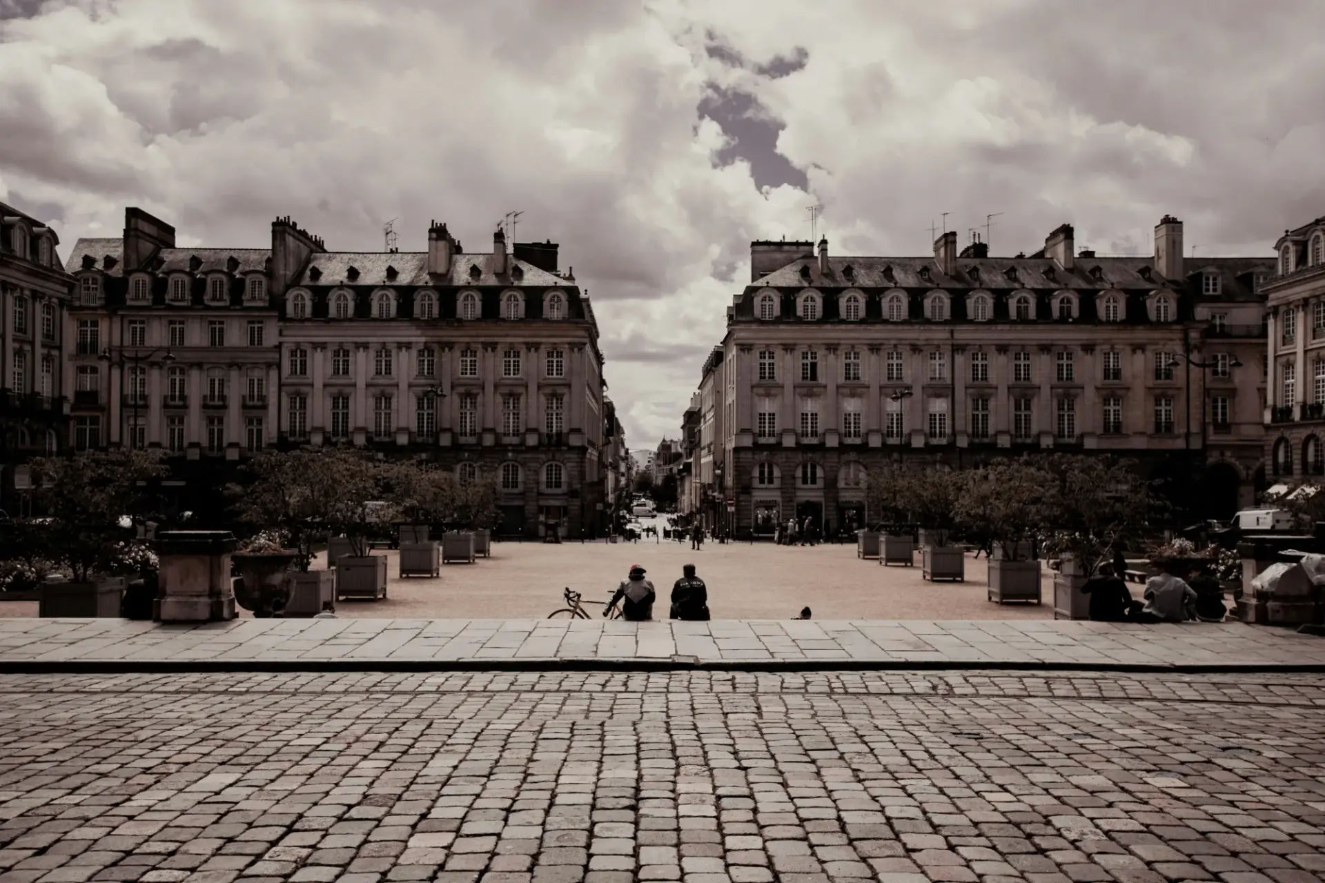 Deux amis assis sur les pavés d'une place du centre de Rennes, face aux façades classiques, ciel nuageux